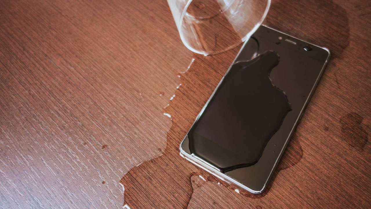 Smartphone on a table covered with water next to a spilled cup of water.