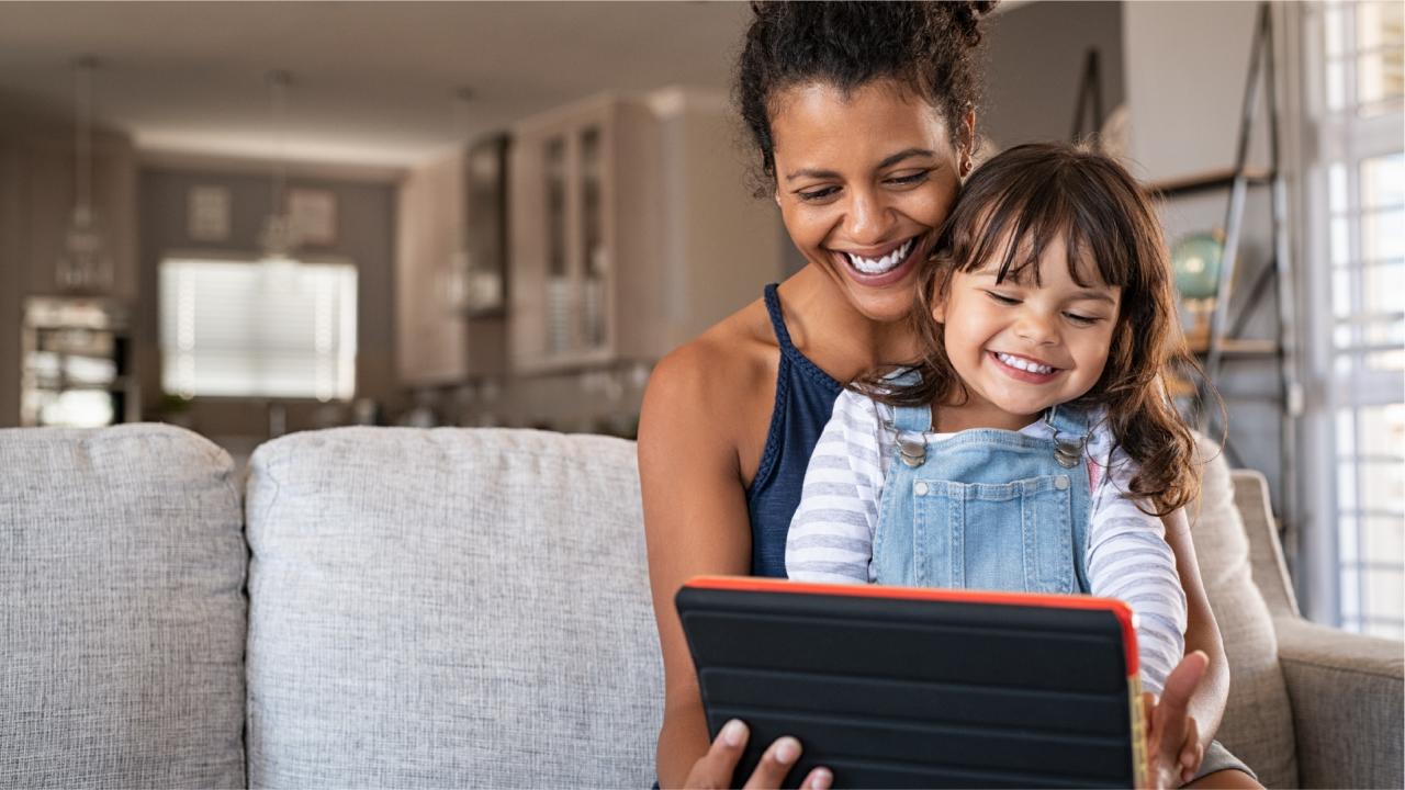 A young child sitting in her mother's lap, smiling and laughing while using a handheld device