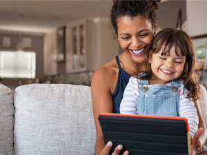 A young child sitting in her mother's lap, smiling and laughing while using a handheld device