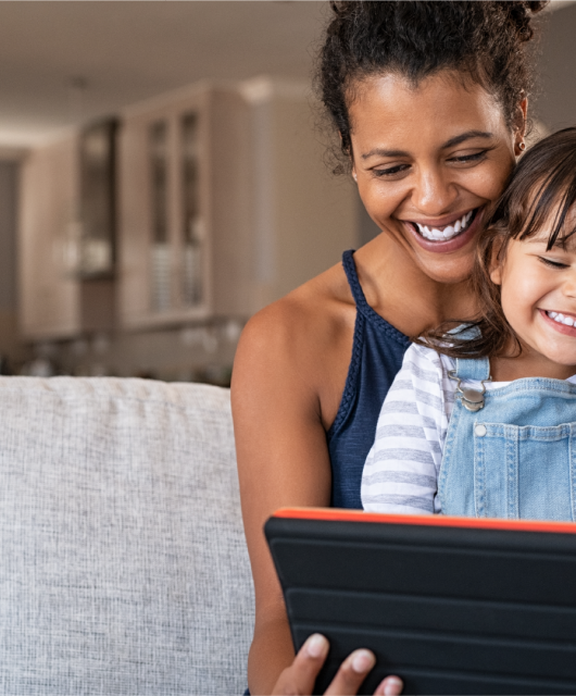 A young child sitting in her mother's lap, smiling and laughing while using a handheld device