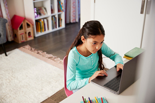 A young girl sits at a desk at home using her laptop computer