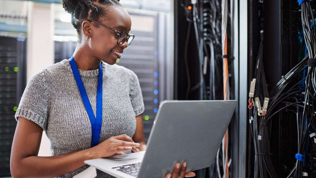 A woman holding a laptop smiles as she looks at a server