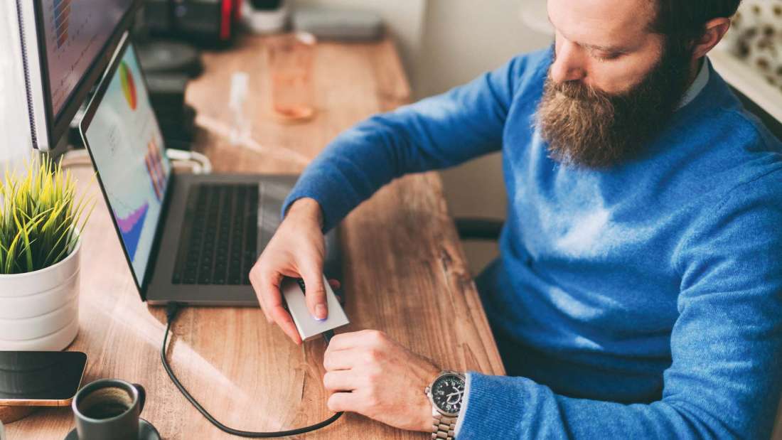 Man in blue sweater with brunette beard plugging external hard drive into laptop.