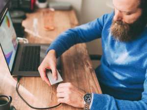 Man in blue sweater with brunette beard plugging external hard drive into laptop.