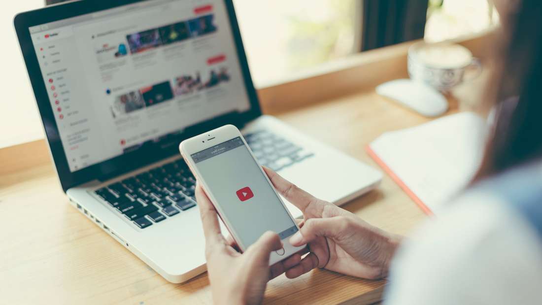 Person loading YouTube on a smartphone with YouTube playlists on a laptop in the background.