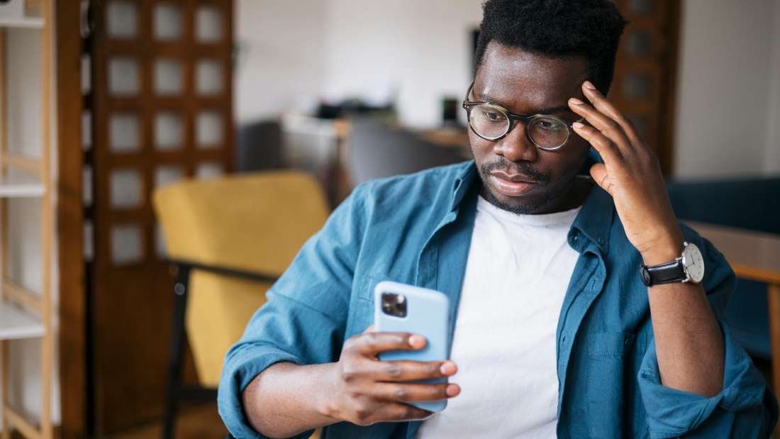 Black man in teal cardigan holding an iPhone with a blue case and looking confused.