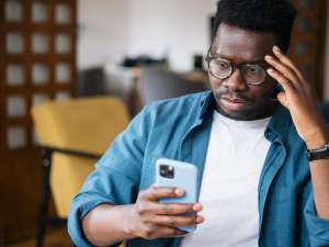 Black man in teal cardigan holding an iPhone with a blue case and looking confused.