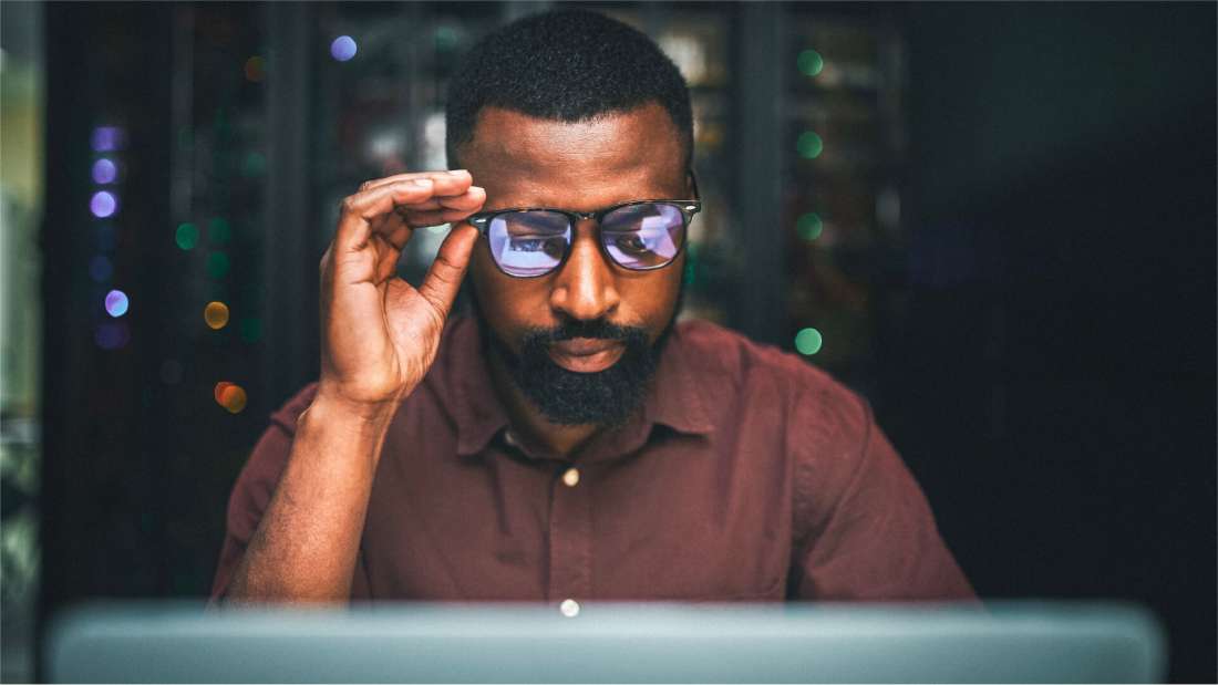 Black man holding his glasses while staring at a computer.