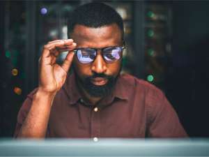 Black man holding his glasses while staring at a computer.