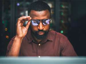 Black man holding his glasses while staring at a computer.