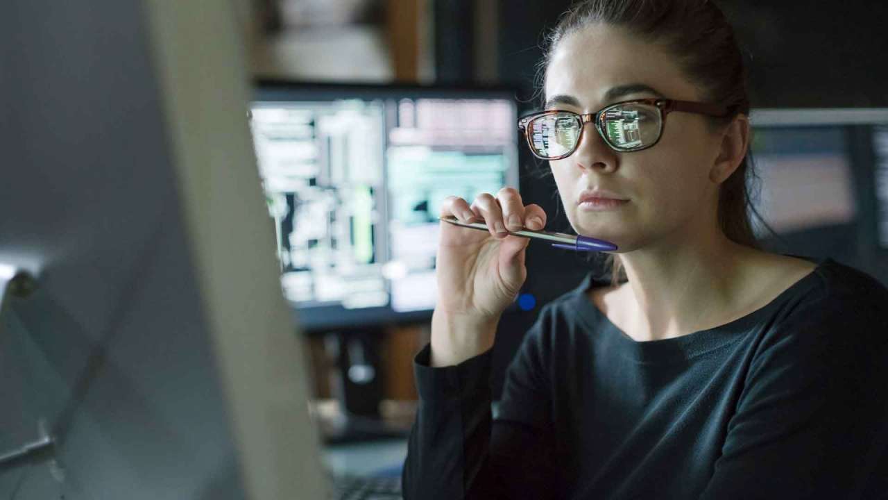 Woman holding pen under her chin looking at a desktop computer.