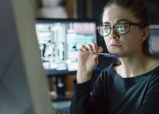 Woman holding pen under her chin looking at a desktop computer.