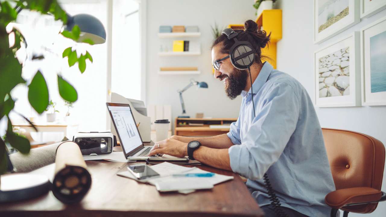 Man wearing headphones and smiling as he types on a laptop in a bright, homey office.