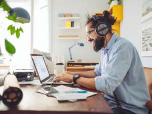 Man wearing headphones and smiling as he types on a laptop in a bright, homey office.