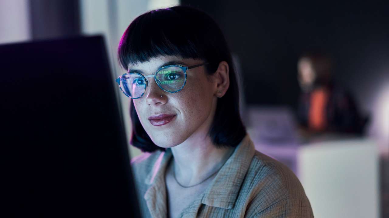 Woman with short dark hair and glasses looking at a lit up computer screen.