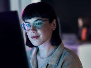 Woman with short dark hair and glasses looking at a lit up computer screen.