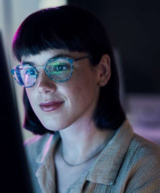 Woman with short dark hair and glasses looking at a lit up computer screen.