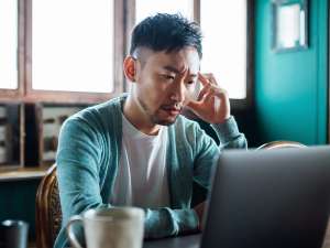 Person sitting in front of a laptop with one hand touching their head, looking stressed.