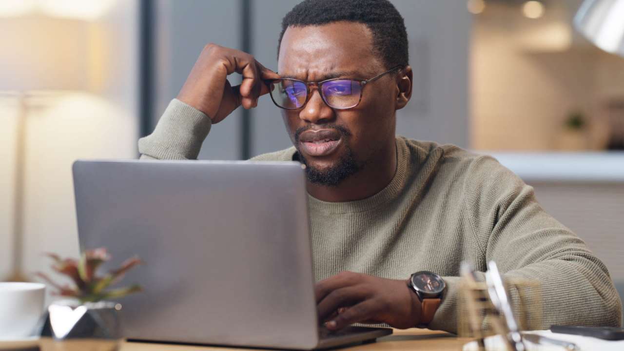 Man holding his glasses on his face while looking confused at a laptop screen.
