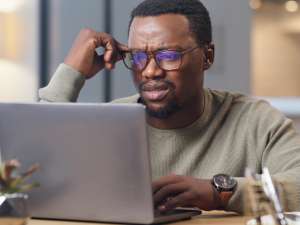 Man holding his glasses on his face while looking confused at a laptop screen.