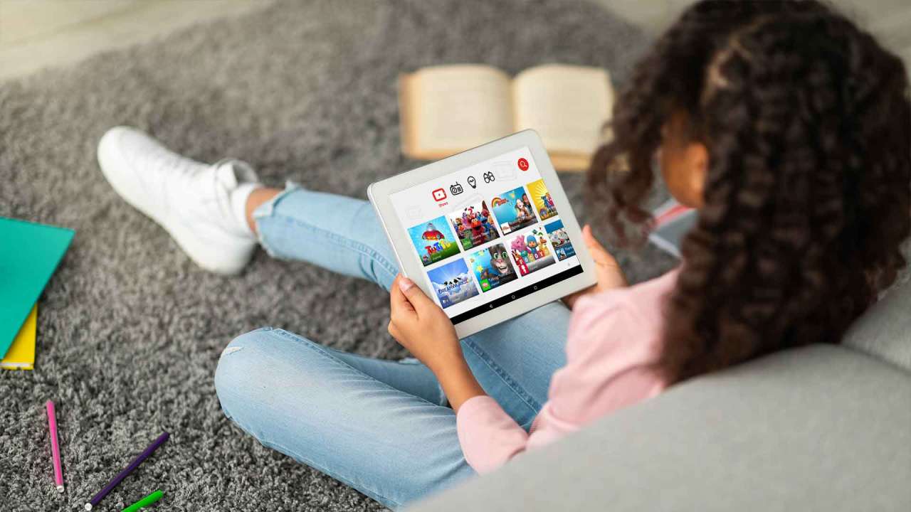 A child sits on the floor of their house against a couch while using YouTube Kids on their tablet