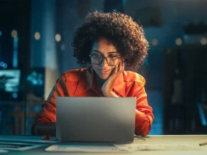 A woman sits at her laptop computer at night, looking intrigued