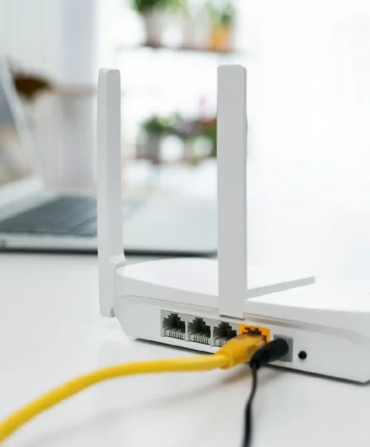 A white colored Wi-Fi router on a table next to a plant and a laptop.
