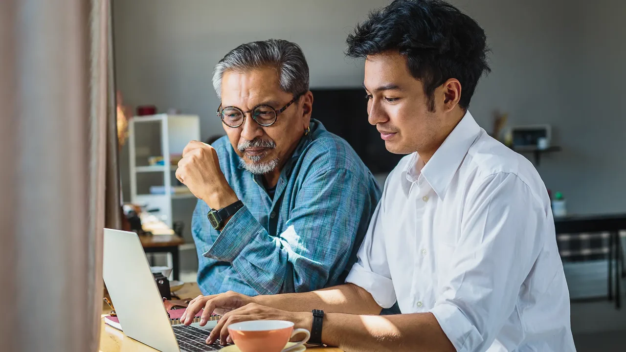 A warm, high-quality lifestyle photograph of a father and son smiling while looking at a laptop together in a bright living room.
