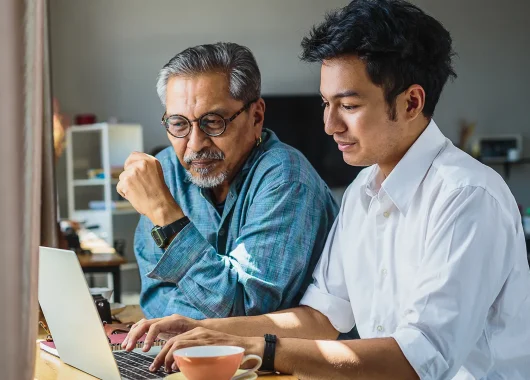 A warm, high-quality lifestyle photograph of a father and son smiling while looking at a laptop together in a bright living room.