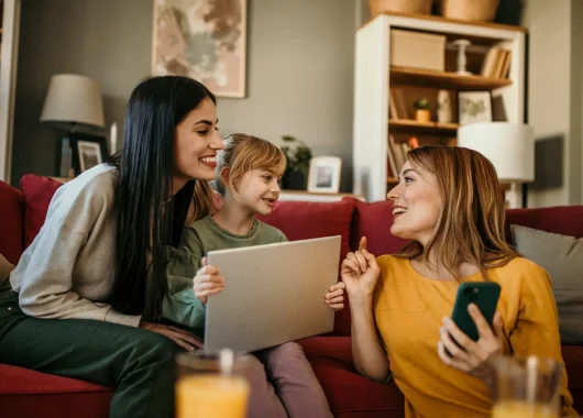 A photo of a family sitting together on a living room couch, comfortably using various devices like a tablet, laptop, and smartphone.