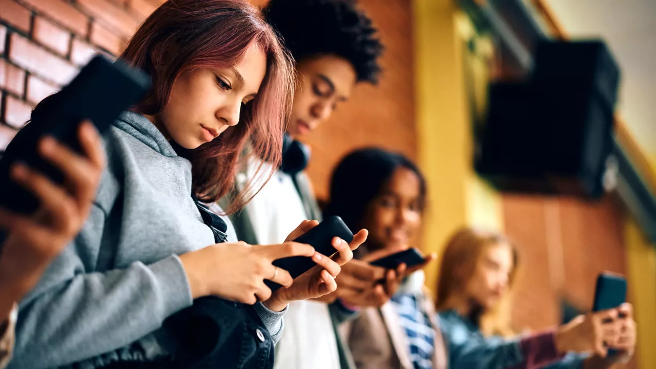 A group of teenagers standing in a row, focused intently on their smartphones.