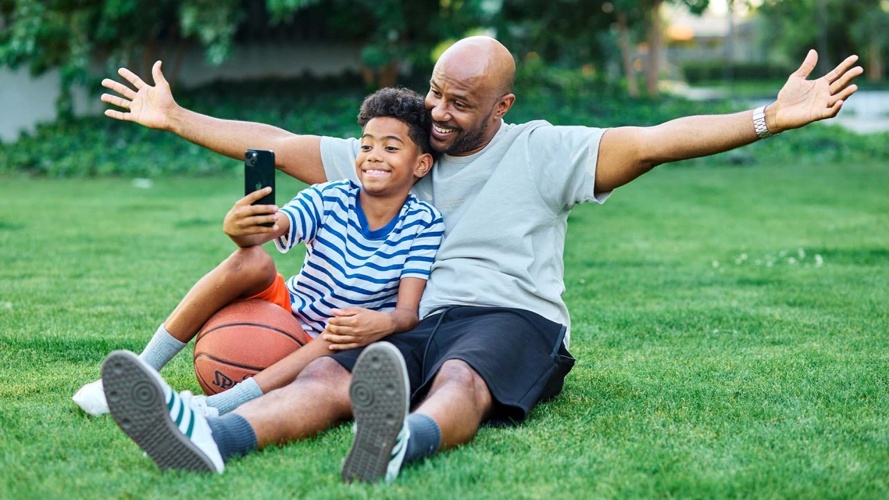 Dad sitting with his son in the grass, taking a selfie together after playing basketball.