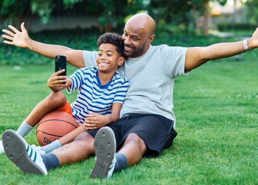Dad sitting with his son in the grass, taking a selfie together after playing basketball.