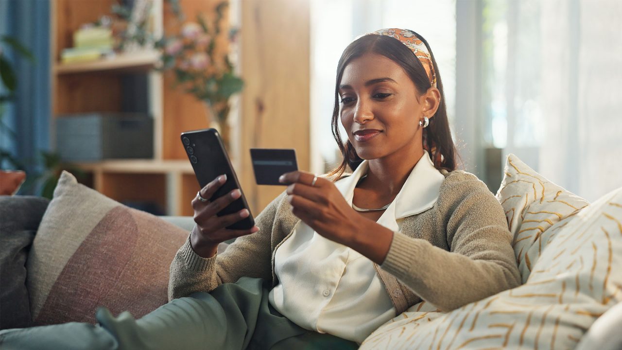 A lady sits on her couch while using her credit card to make an online purchase.