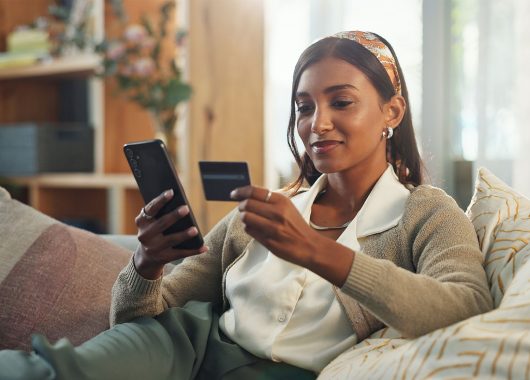 A lady sits on her couch while using her credit card to make an online purchase.