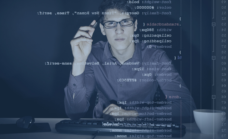 young man with glasses sitting in front of his computer, programming. the code he is working on (CSS) can be seen through the screen.